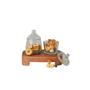 Two glass jars with a vintage design, one larger and one smaller, placed on a wooden tray. The larger jar has a lid with a rounded knob.